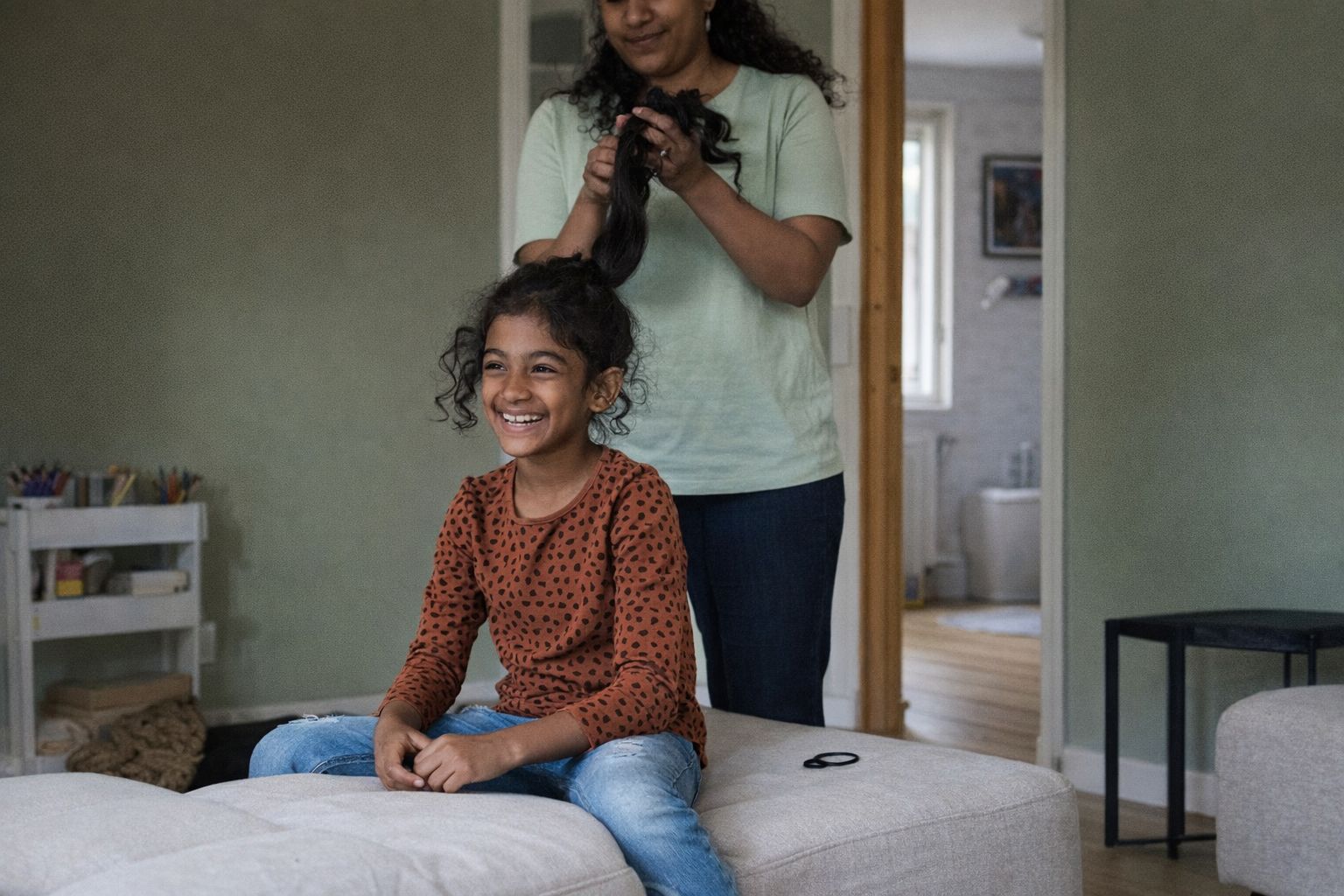 En leende flicka får sitt hår borstat av en vuxen i ett hemtrevligt vardagsrum.
