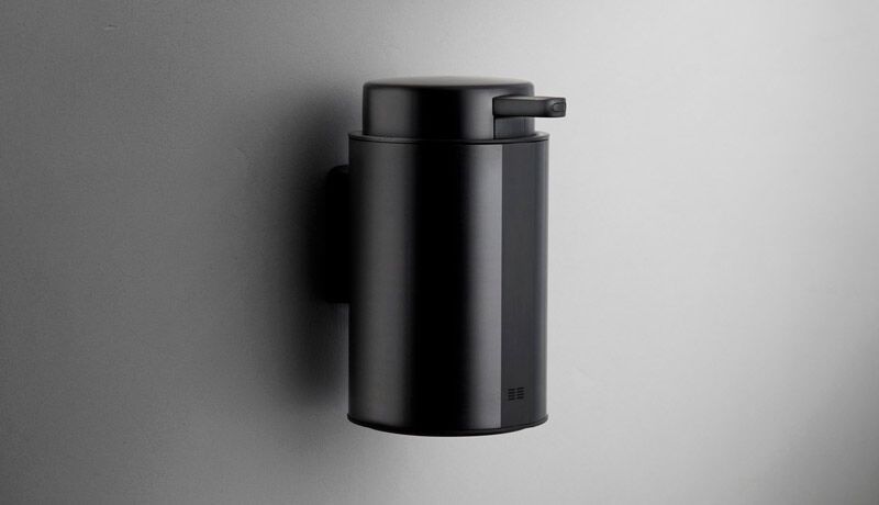 Väggmonterad svartvålpump i minimalistisk design på grå vägg.